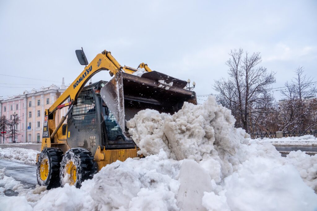 A yellow tractor clears snow on a city street after a winter snowstorm, showcasing urban cleanup.